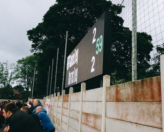 A large digital scoreboard at an outdoor sports venue displays a score of 2-2. Spectators are gathered, observing the event with interest. The scene is enclosed by a tall metal fence and surrounded by lush green trees, indicating a natural setting. Overcast skies add a muted tone to the environment.