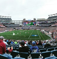 A large sports stadium filled with spectators. The field is green with white markings, and players in red and white uniforms are actively engaged in a game. The stands are packed with people wearing various colored clothing, and some sections of the crowd are more densely populated. Digital screens and advertisements are visible around the stadium, adding to the vibrant atmosphere.