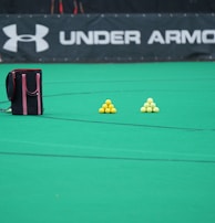 A sports field features a black and pink sports bag, two pyramids of yellow balls, and a portion of a person in dark athletic gear. The background displays a banner with the Under Armour logo.