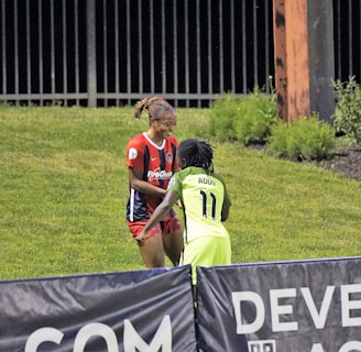 Two individuals are interacting on a grassy field. One is wearing a red and blue sports outfit with 'ProChain' visible, and the other is in a bright yellow uniform with the name 'Addo' and the number 11. They appear to be playing a sport, possibly soccer, as they are near a marked boundary. A fence and some vegetation are visible in the background.