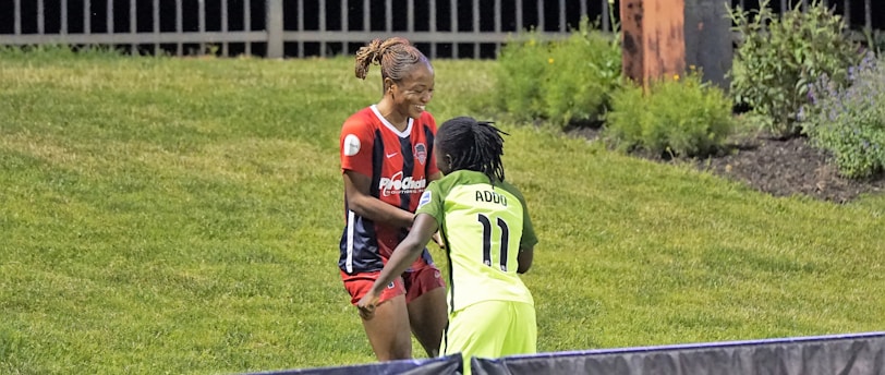 Two individuals are interacting on a grassy field. One is wearing a red and blue sports outfit with 'ProChain' visible, and the other is in a bright yellow uniform with the name 'Addo' and the number 11. They appear to be playing a sport, possibly soccer, as they are near a marked boundary. A fence and some vegetation are visible in the background.