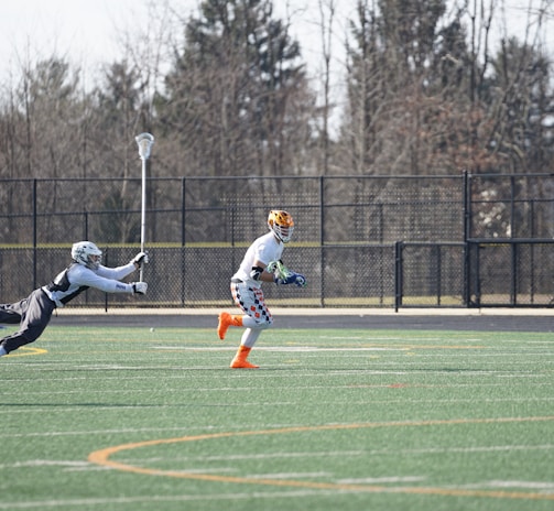 A group of athletes engaged in a sports game on a green field. One player is running forward with possession of the ball, wearing colorful socks and a checkered uniform, while another player in dark clothing dives to intercept or tackle. Other players are present in the background. Leafless trees and a metal fence surround the field.