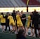A group of athletes wearing yellow sports uniforms are standing on a sports field. Some are holding lacrosse sticks, and they appear to be interacting casually. Several people are dressed in black coats. In the background, there is a bright sign with text and parts of a stadium are visible.