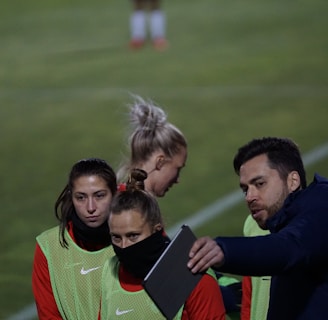 A group of individuals on a sports field are wearing green vests over red attire. One person is holding a tablet and seems to be discussing or showing something to the others. The atmosphere suggests a sports event, likely soccer, indicated by their clothing and the setting. The background is a grassy field.