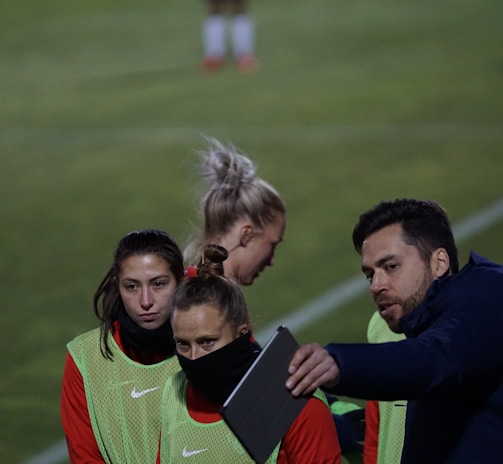 A group of individuals on a sports field are wearing green vests over red attire. One person is holding a tablet and seems to be discussing or showing something to the others. The atmosphere suggests a sports event, likely soccer, indicated by their clothing and the setting. The background is a grassy field.