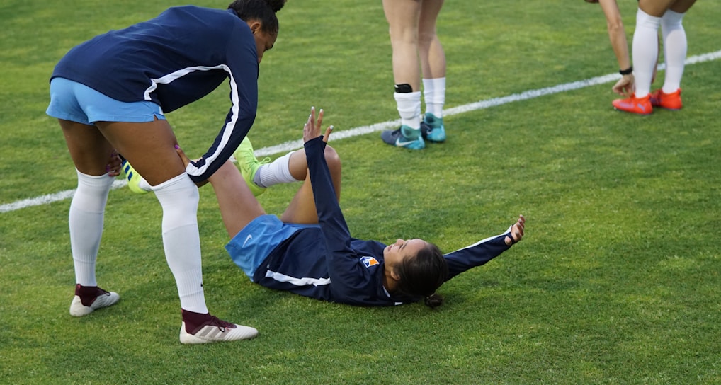 A group of athletes in sportswear are on a green soccer field. Two individuals are interacting closely; one helps the other who is lying on the grass. Nearby, other athletes are stretching.