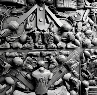 A complex arrangement of various sports equipment and attire, including tennis rackets, soccer balls, basketballs, helmets, shoes, gloves, and a mannequin torso wearing a T-shirt. The items are layered and artistically displayed in a monochrome color scheme, creating an intricate texture and visual pattern.
