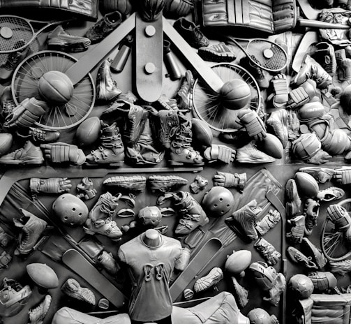 A complex arrangement of various sports equipment and attire, including tennis rackets, soccer balls, basketballs, helmets, shoes, gloves, and a mannequin torso wearing a T-shirt. The items are layered and artistically displayed in a monochrome color scheme, creating an intricate texture and visual pattern.