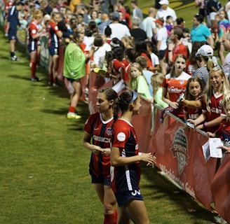 The image features a lively gathering of young soccer players and fans next to a field. Several female athletes in red and blue uniforms interact with an enthusiastic crowd standing behind a barrier. Some people in the crowd are holding objects, like pens and paper, possibly for autographs.