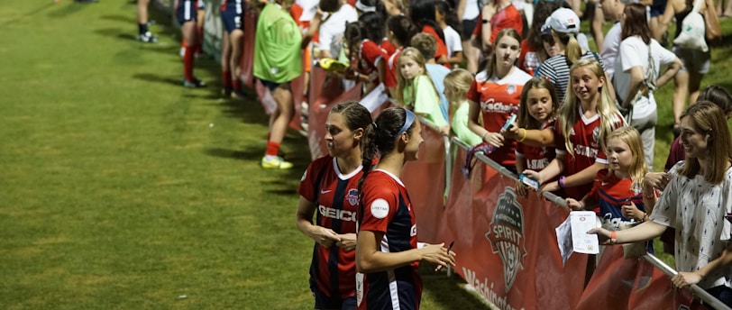 The image features a lively gathering of young soccer players and fans next to a field. Several female athletes in red and blue uniforms interact with an enthusiastic crowd standing behind a barrier. Some people in the crowd are holding objects, like pens and paper, possibly for autographs.