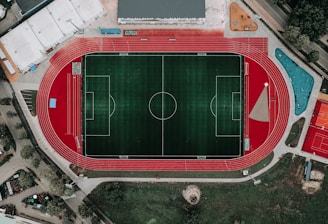 An aerial view of a large sports complex featuring a central green football field surrounded by a red running track. Adjacent to the track, there is a parking area, buildings, and pathways. Nearby, there are additional sports courts in different colors, including a blue swimming pool and orange basketball courts.