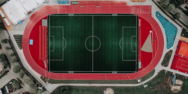 An aerial view of a large sports complex featuring a central green football field surrounded by a red running track. Adjacent to the track, there is a parking area, buildings, and pathways. Nearby, there are additional sports courts in different colors, including a blue swimming pool and orange basketball courts.