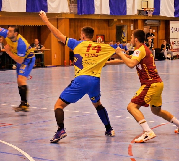 A dynamic indoor sports scene with players in motion on a court. Two athletes in blue and yellow uniforms are engaged in an intense handball game, with one player gripping the other's jersey. The background shows other players, several seated people, and advertisements on the walls.