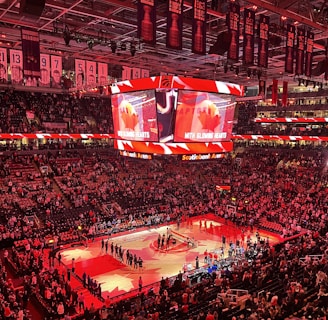An indoor sports arena filled with spectators and a basketball court illuminated by red lighting. The scoreboards and video screens display promotional content, with numerous banners and championship flags hanging from the ceiling. Players and officials are gathered on the court, possibly in preparation for a game or event.