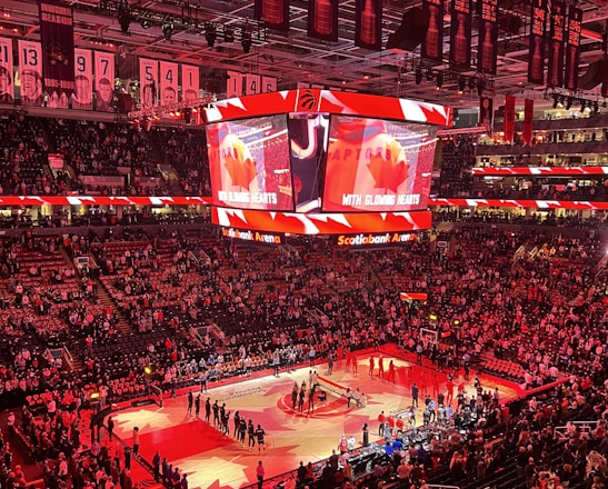 An indoor sports arena filled with spectators and a basketball court illuminated by red lighting. The scoreboards and video screens display promotional content, with numerous banners and championship flags hanging from the ceiling. Players and officials are gathered on the court, possibly in preparation for a game or event.