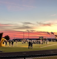 A sports field with a football game unfolding under a vibrant sunset sky, featuring a large inflatable mascot and a scoreboard displaying the score. Players and coaches are visible on the green field, surrounded by a track and some flags waving in the breeze.