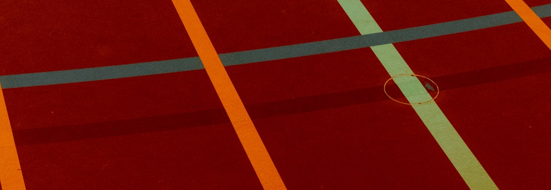 A sports court with intersecting lines in orange and blue, creating geometric patterns on a reddish-brown surface.