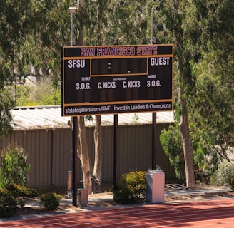 An outdoor scoreboard for San Francisco State is seen among trees and greenery. The board displays sections for shots on goal and corner kicks, with areas for both home and guest teams. It is situated near a running track with a building in the background.