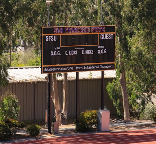 An outdoor scoreboard for San Francisco State is seen among trees and greenery. The board displays sections for shots on goal and corner kicks, with areas for both home and guest teams. It is situated near a running track with a building in the background.