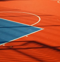 A vibrant sports court with sections of red and blue, featuring crisp white lines marking the boundaries. Shadows from a structure create geometric patterns across the surface, adding depth and interest to the scene.
