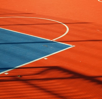 A vibrant sports court with sections of red and blue, featuring crisp white lines marking the boundaries. Shadows from a structure create geometric patterns across the surface, adding depth and interest to the scene.
